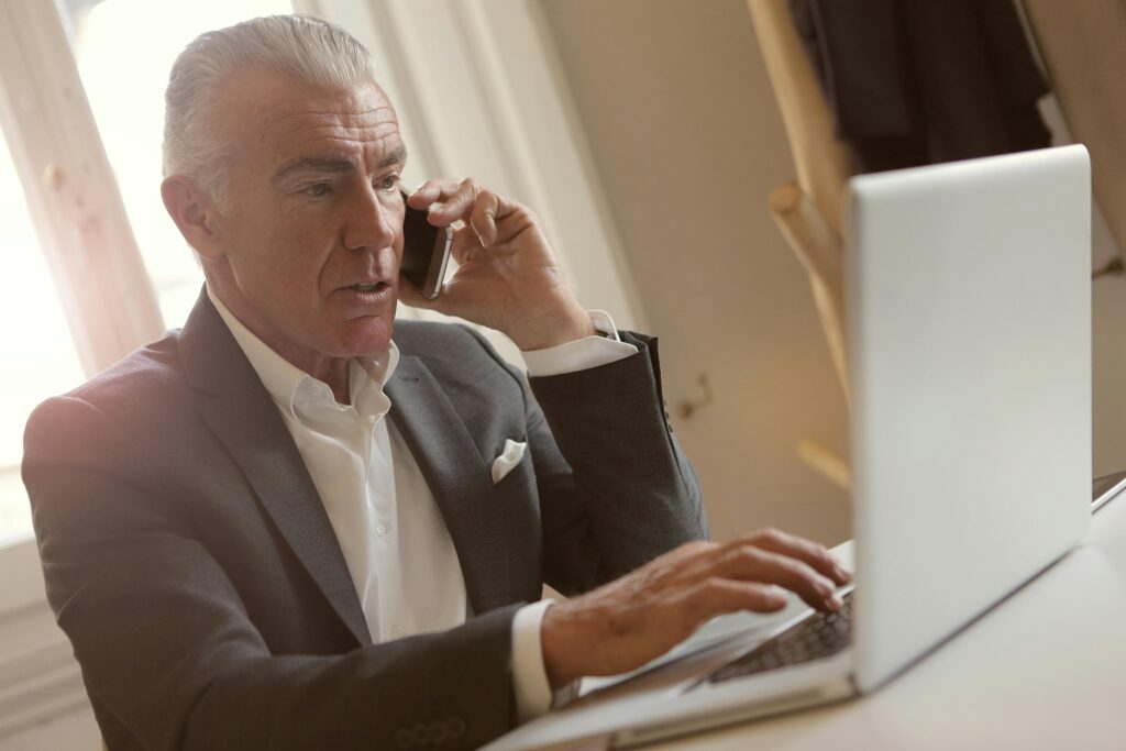 Senior businessman in a suit discussing business sale on a phone call while working on a laptop.